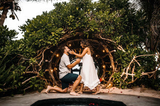A man proposing to his girlfriend on one knee with a beautiful green jungle backdrop and fairy lights