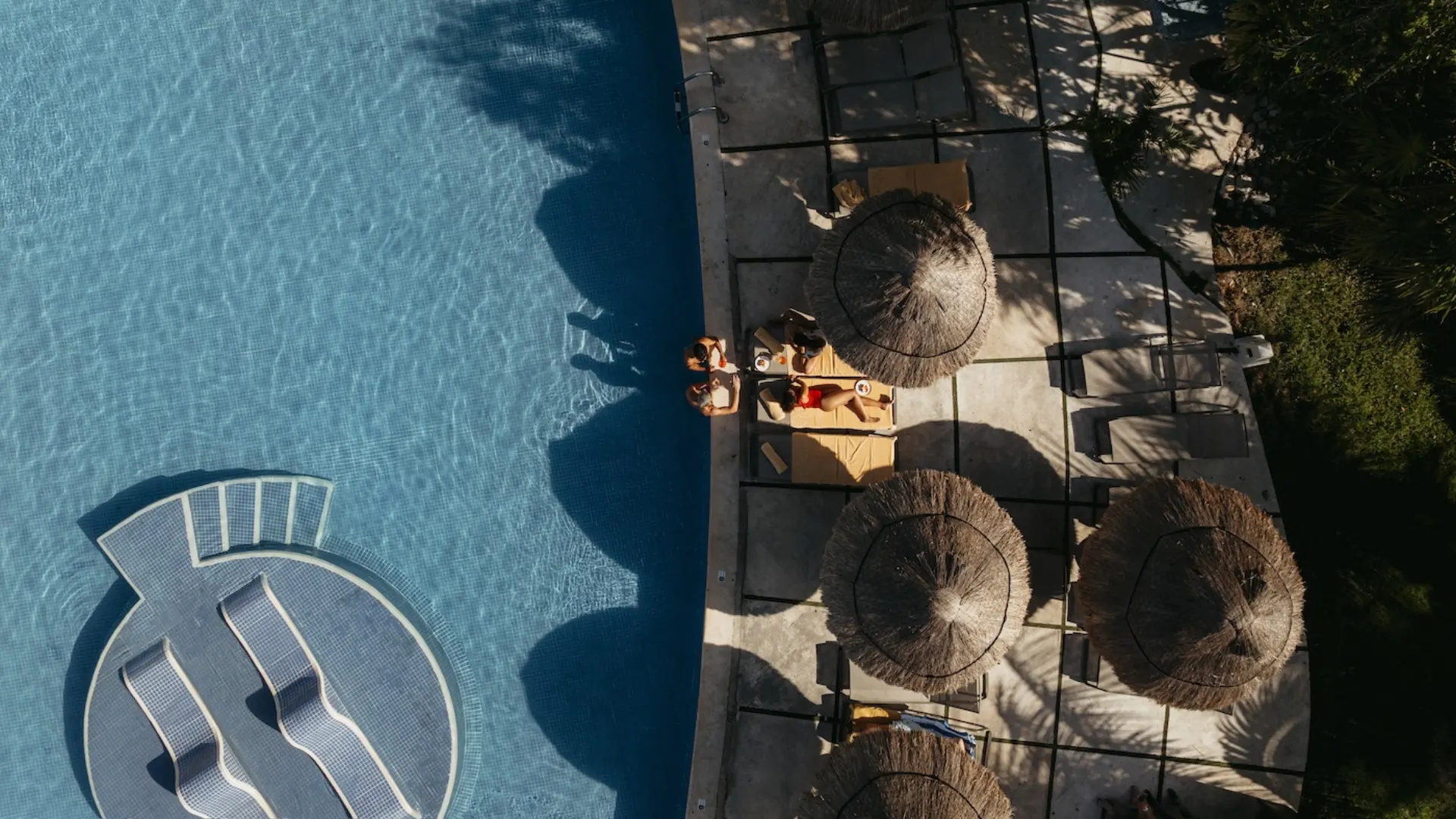 Aerial view of guests relaxing by a resort pool under thatched palapa umbrellas