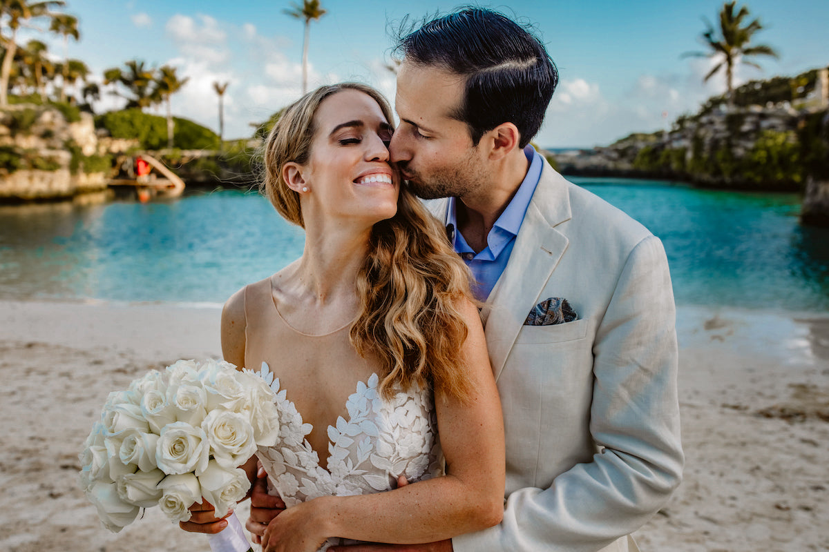 Groom kisses his smiling bride's cheek beside a turquoise lagoon lined with palm trees