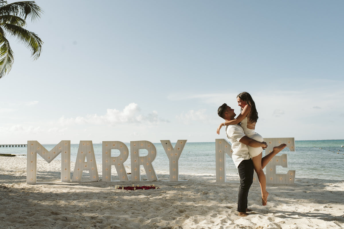 Man lifts his fiancee before a lit MARRY ME sign on the beach after a surprise proposal