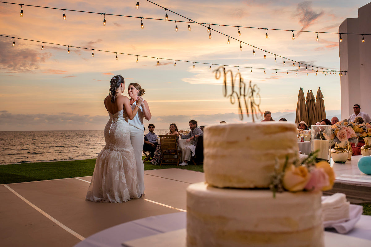 Two brides share their first dance at a seaside reception under string lights at sunset