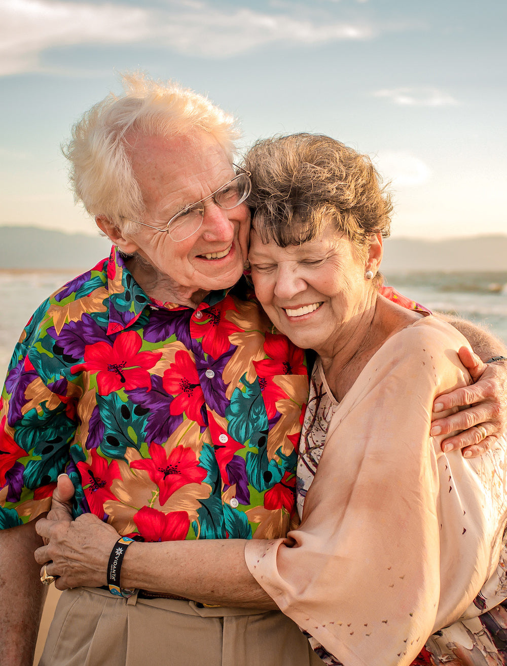 Elderly couple cuddles cheek to cheek on the beach at sunset, husband in a bright floral shirt