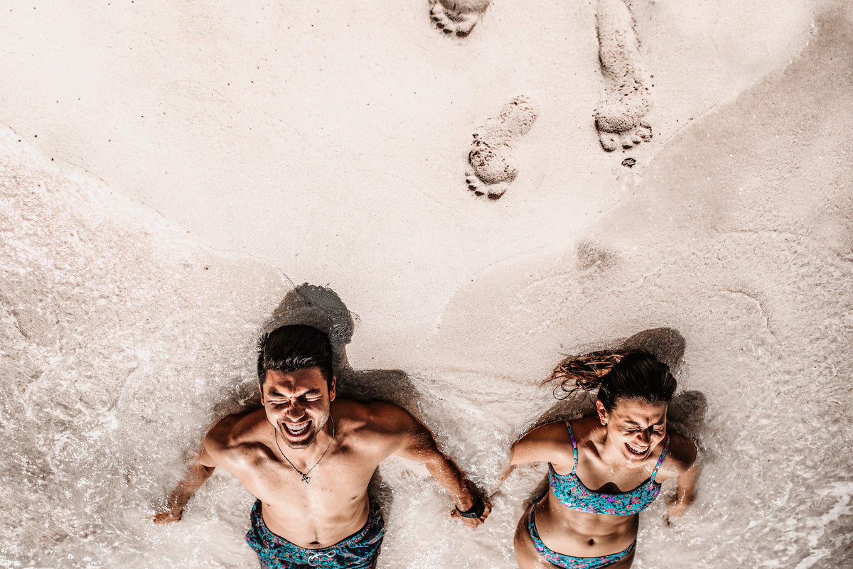 Laughing couple lying in the white sand and surf seen from above during a couples photoshoot