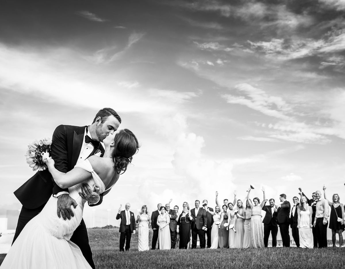 Groom dips and kisses his bride on a lawn as cheering wedding guests raise their arms behind them