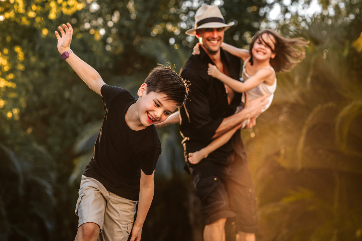Dad in a fedora swings his daughter while his son plays airplane in a sunlit garden family photoshoot