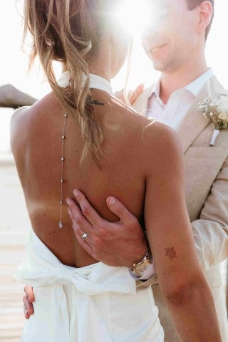 Photograph of groom holding the bride. There is a focus on her drop-back necklace and his ring.