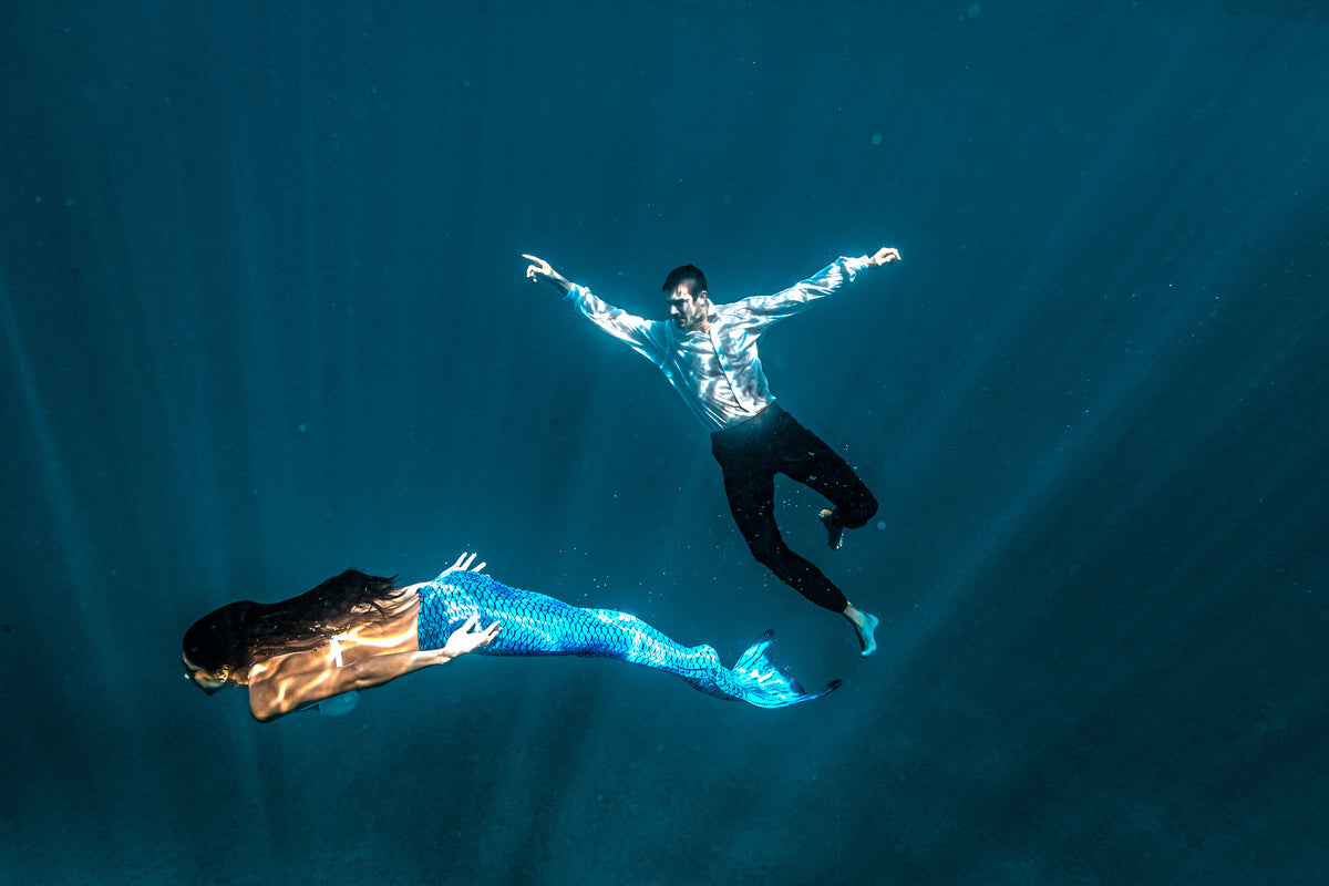 Man in a white shirt and a woman with a blue mermaid tail swim underwater through sunbeams