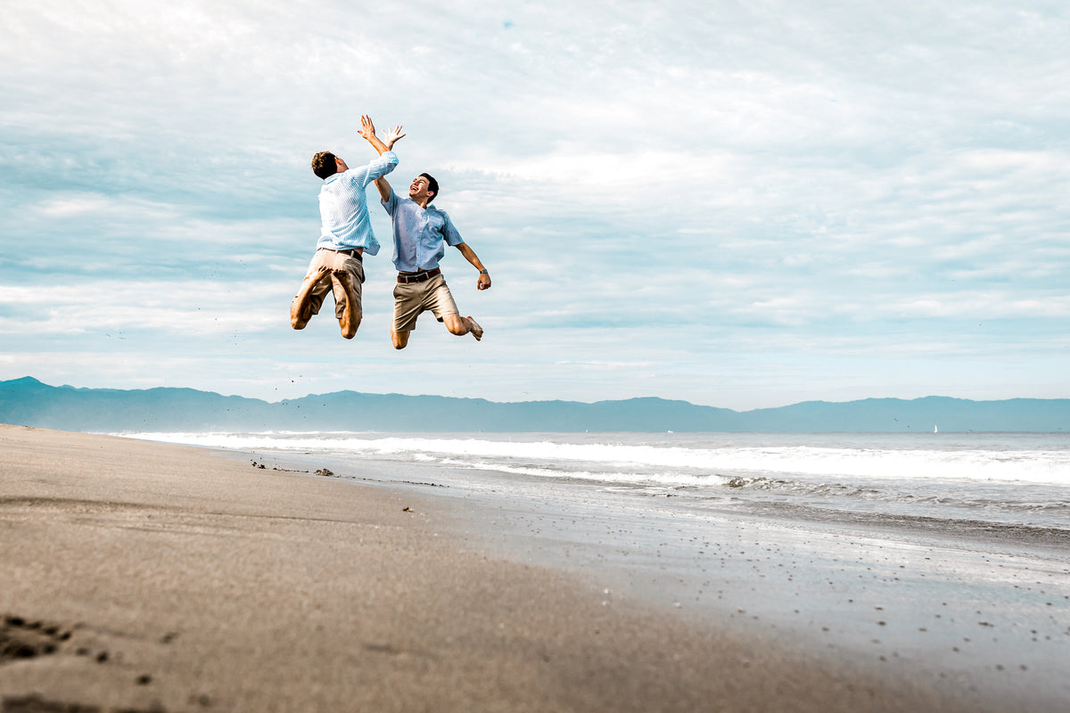 Two men jump for a mid-air high-five on the beach in Vallarta with mountains behind