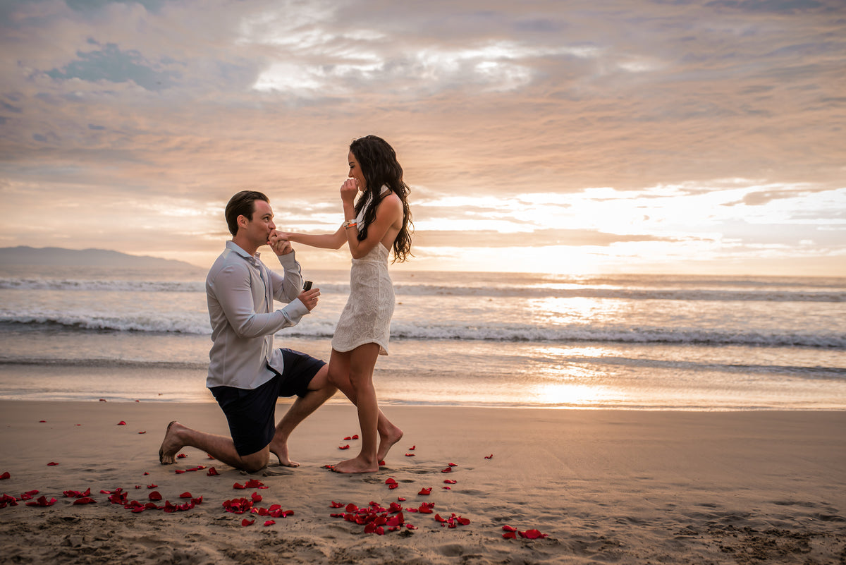 Man kneels to propose to his girlfriend on a rose petal-covered beach at sunset