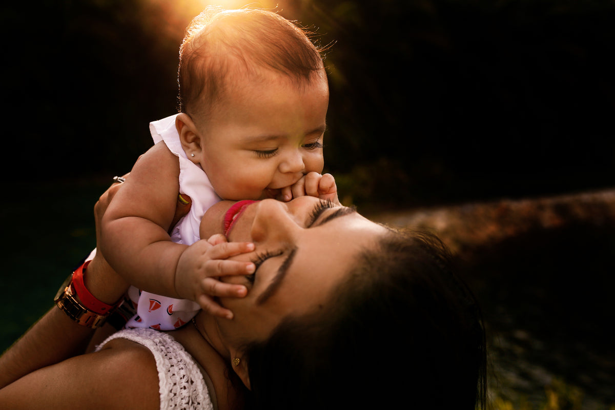 Mother kisses her baby at golden hour with the sun glowing behind them during a family photoshoot