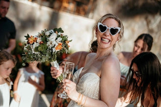 Photograph of a bride holding flowers, champagne, and wearing silly heart-shaped sun glasses. Captured by a DreamArt photographer