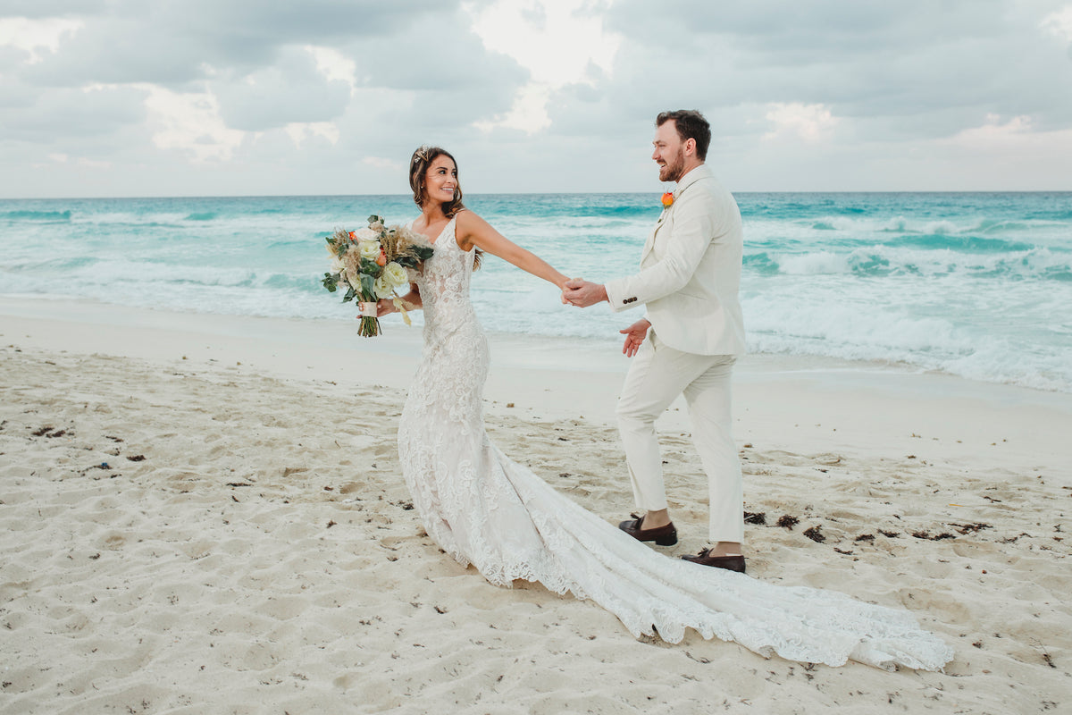 Bride in a lace gown with a long train leads the groom by the hand along a turquoise beach