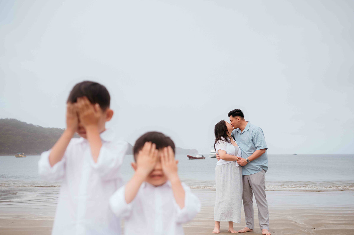 Family photograph of two children covering their eyes whilst their parents kiss behind them on the beach, captured by a DreamArt photographer