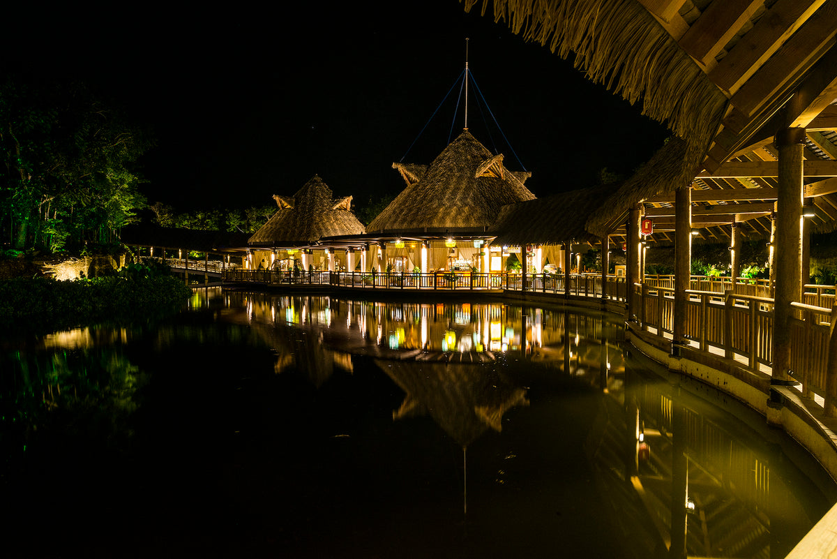 Thatched-roof restaurant pavilions lit at night and reflected in a dark lagoon, architecture photography