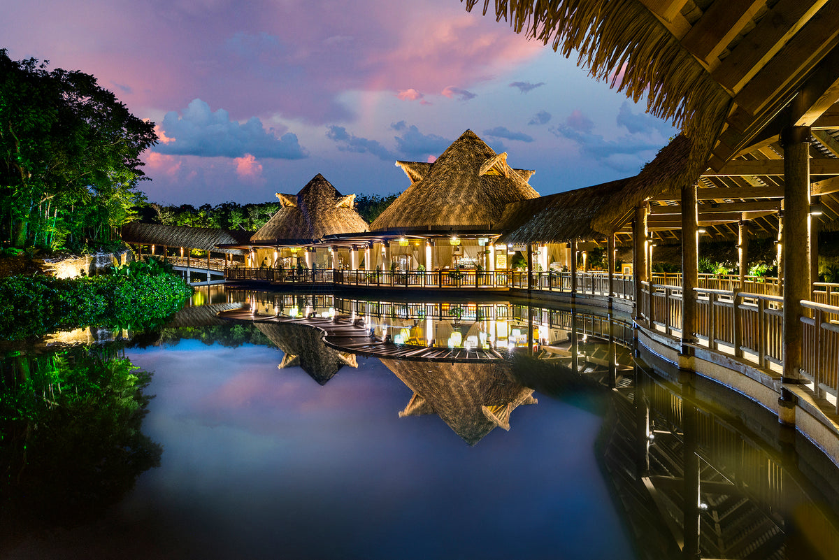Thatched pavilions glowing at dusk under a pink sky, mirrored in a still lagoon, resort architecture photography