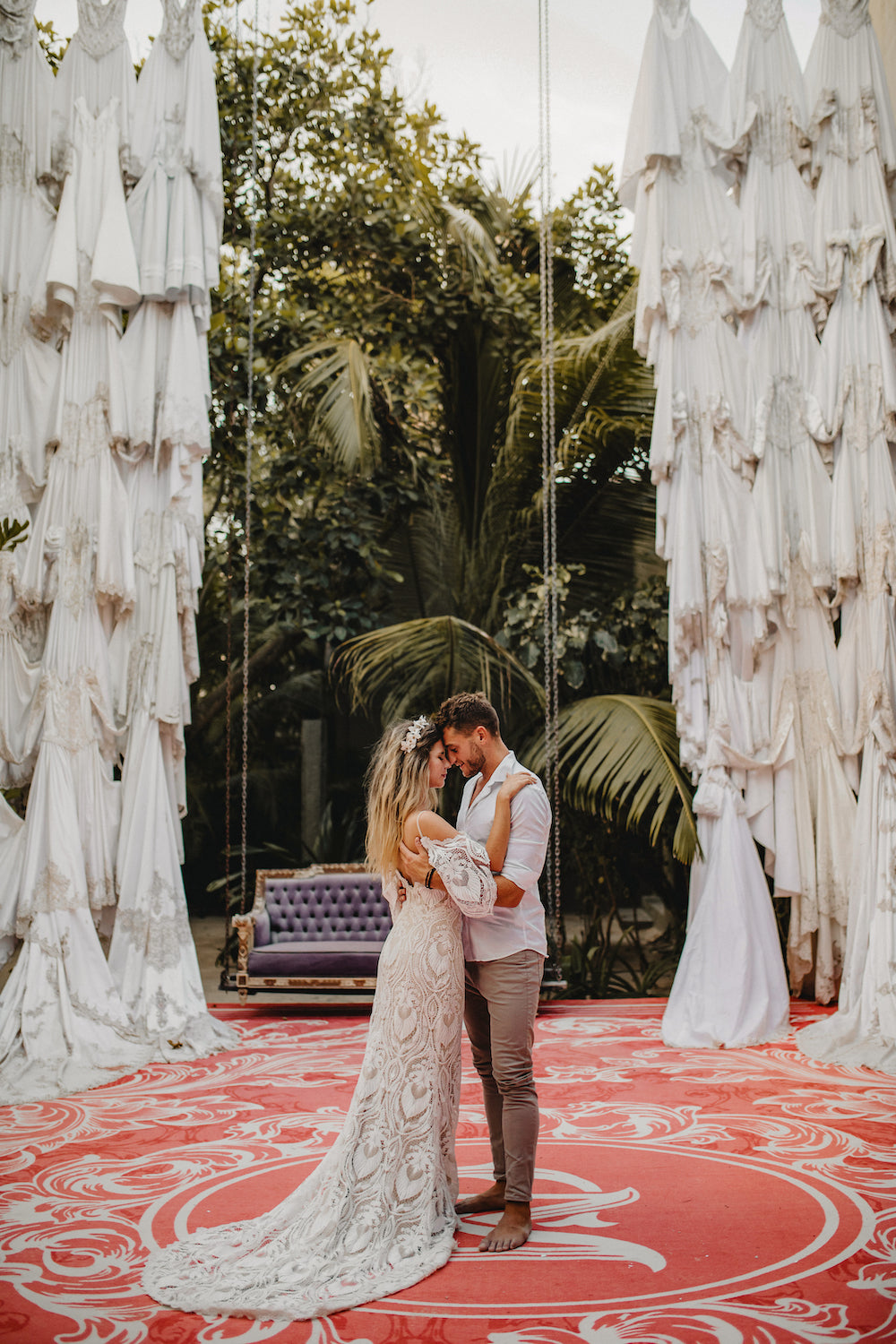 Couple embrace forehead to forehead on a red patterned floor framed by hanging white dresses at Casa Malca Tulum