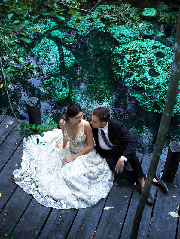 Bride in a lace gown and groom sit on a wooden deck above a green cenote