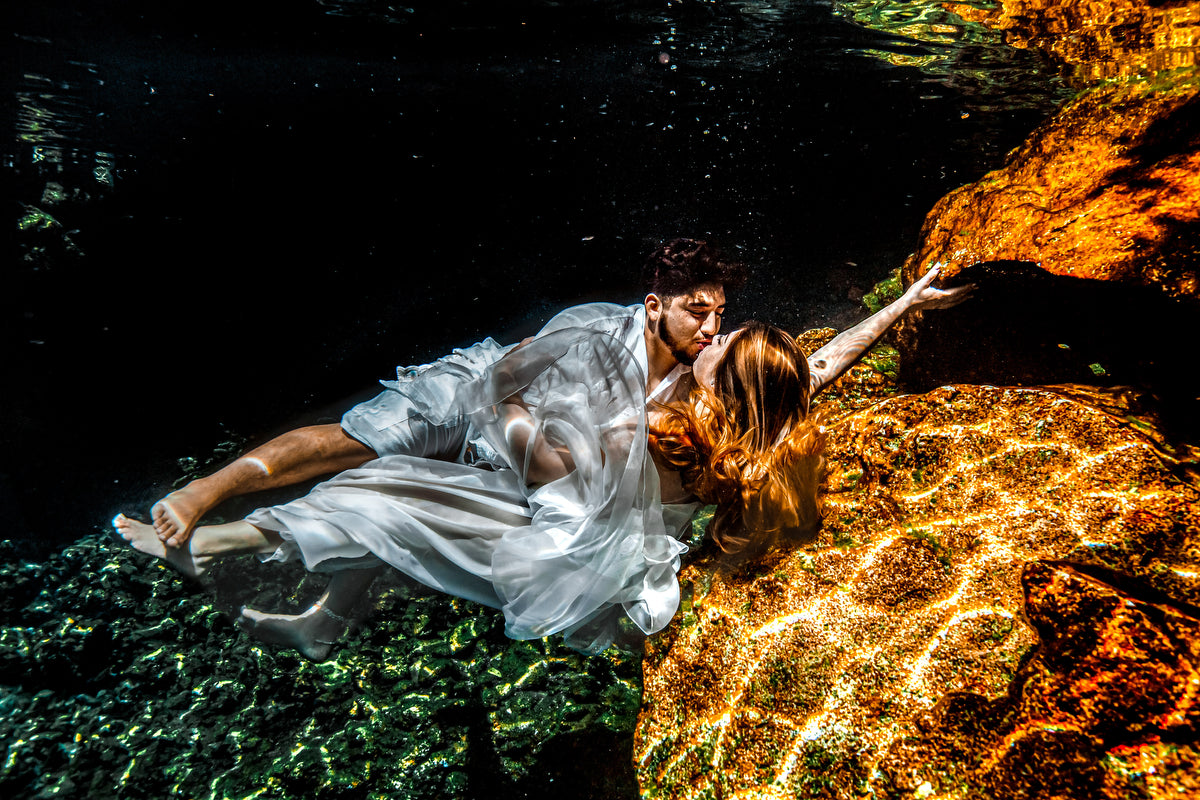 Couple in white kisses underwater over sunlit rocks in a cenote during a trash the dress photoshoot