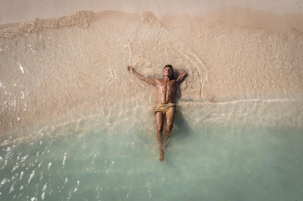 Drone view of a man floating in clear shallow water at the edge of a white sand beach