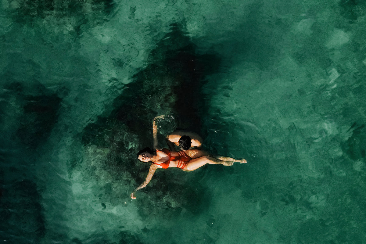 Drone view of a couple floating together in emerald sea water, the woman in a red swimsuit