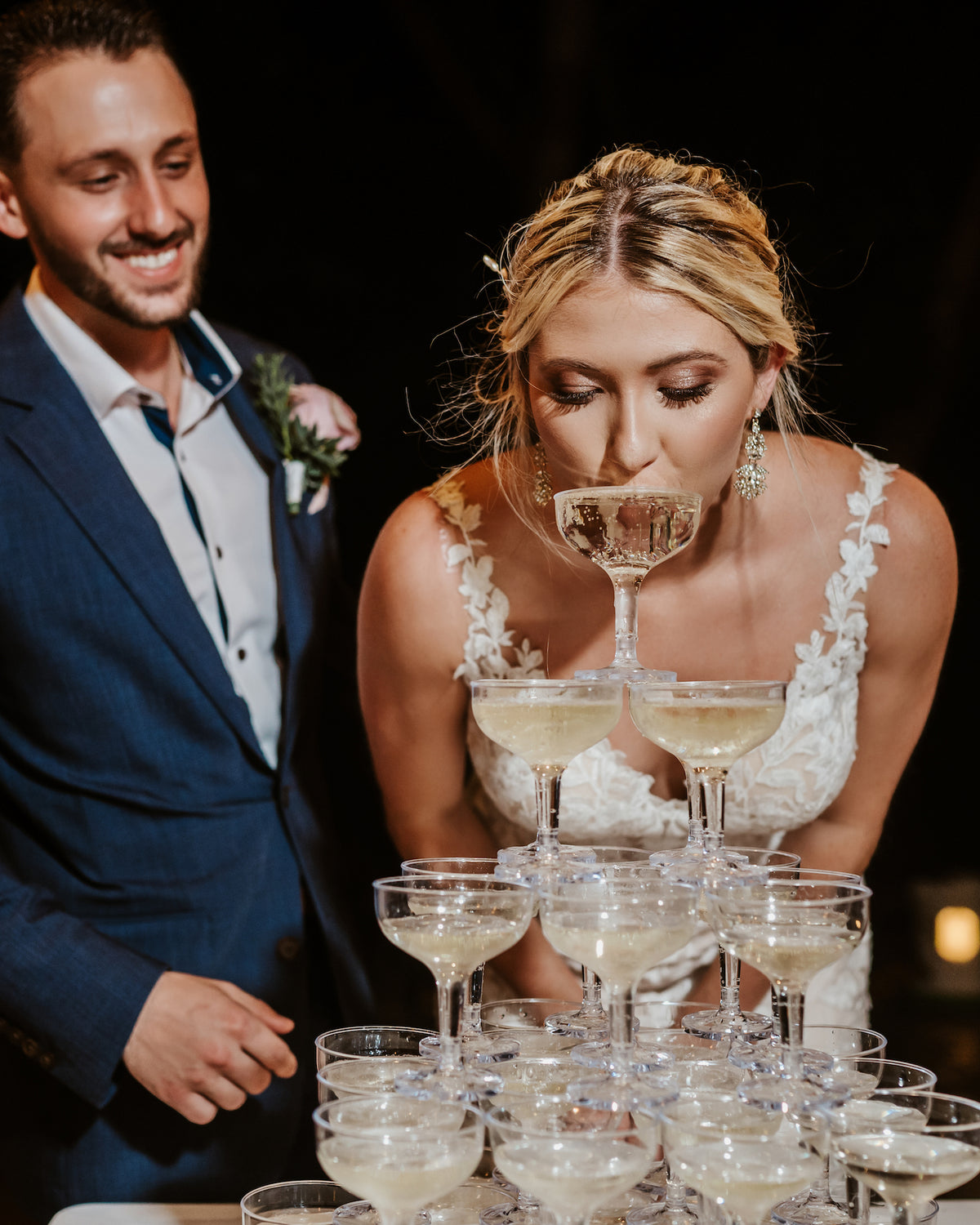 Bride sips from the top of a champagne tower as the groom smiles at their wedding reception