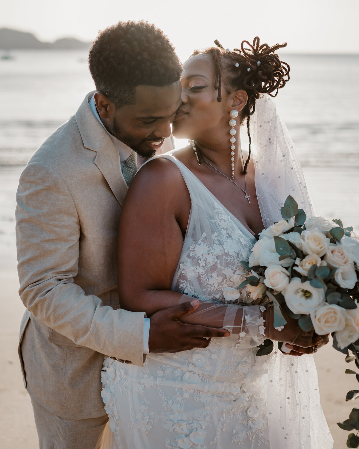 Groom hugs his bride from behind as she kisses his cheek during a beach wedding photoshoot