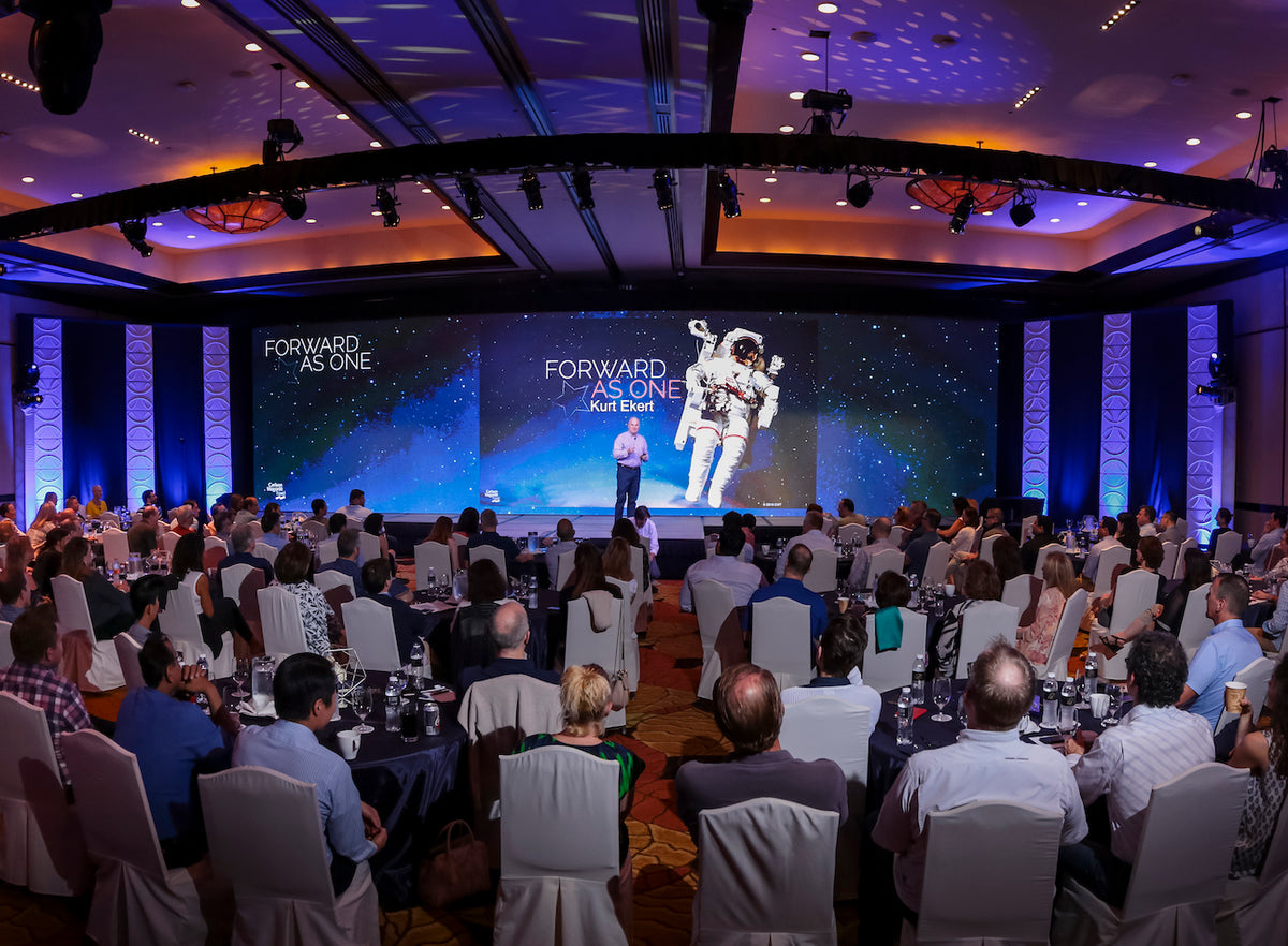 A speaker presents on stage before a seated audience with a large screen reading "Forward As One