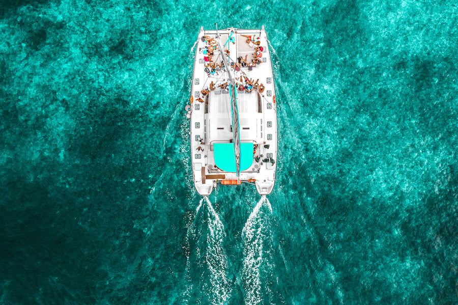 Aerial view of a white catamaran with people aboard, leaving twin wakes in turquoise water