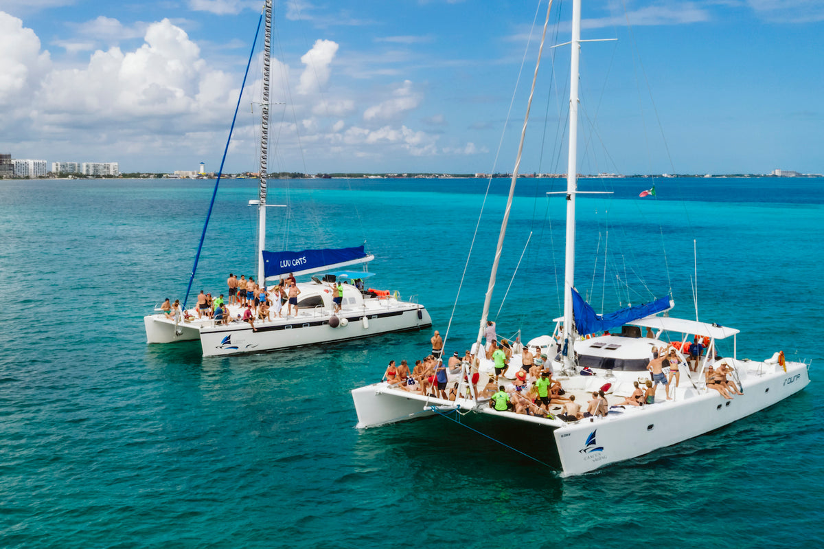 Two catamarans with passengers gathered on deck float on turquoise water near a coastal skyline