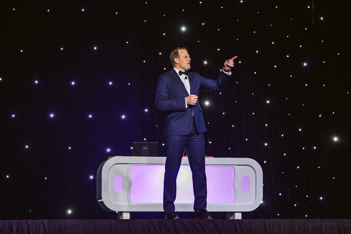 A man in a navy suit points upward while speaking on stage with a starlit backdrop