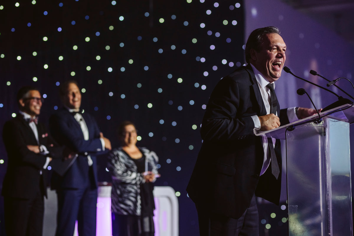 A man in a suit speaks animatedly at a clear podium with microphones on an event stage