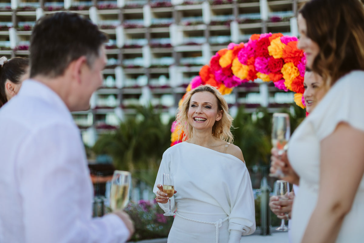 Smiling guest in a white off-shoulder dress raising champagne under a bright flower arch at an event cocktail