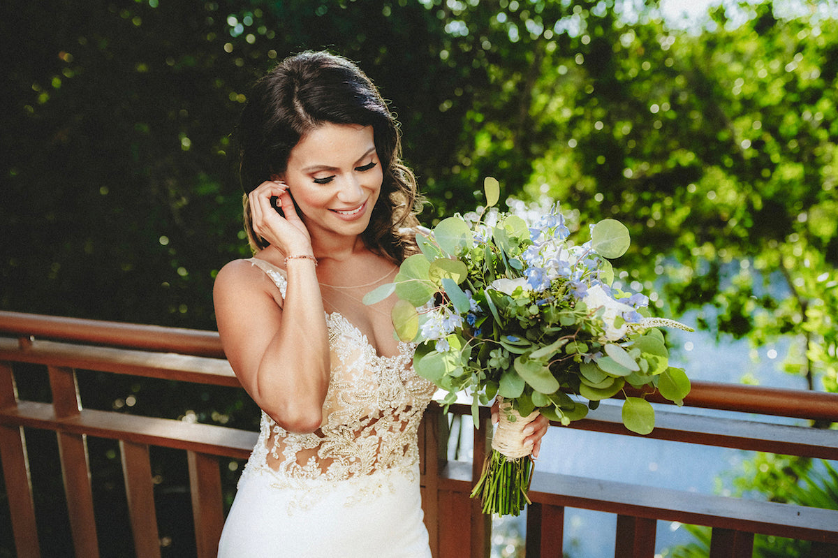 Smiling bride in a lace gown holds a eucalyptus and hydrangea bouquet on a wooden deck at Fairmont Mayakoba