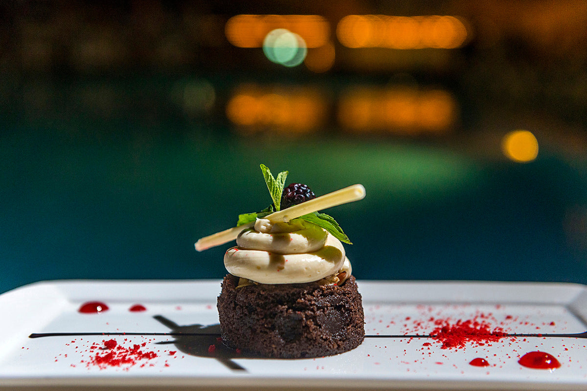 Chocolate dessert topped with cream, mint and a blackberry on a white plate beside a lit pool at night