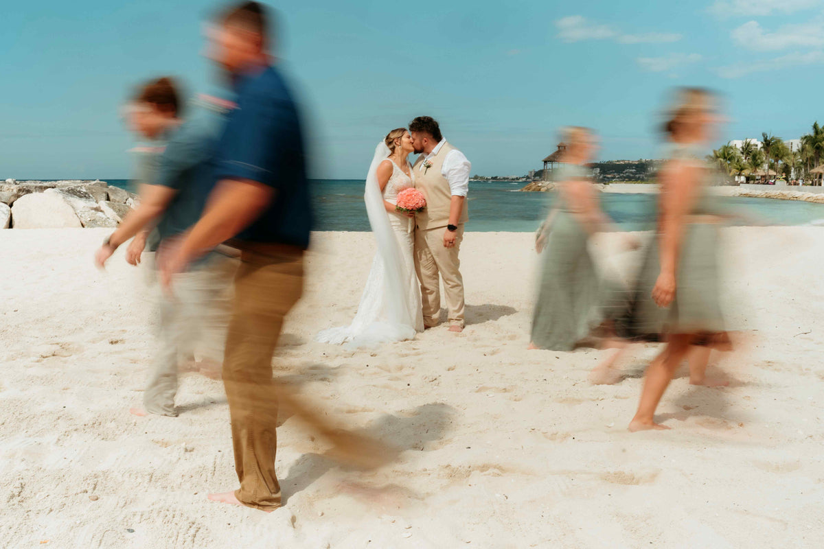 Artistic photograph of bride and groom kissing on the beach as guests walk past them