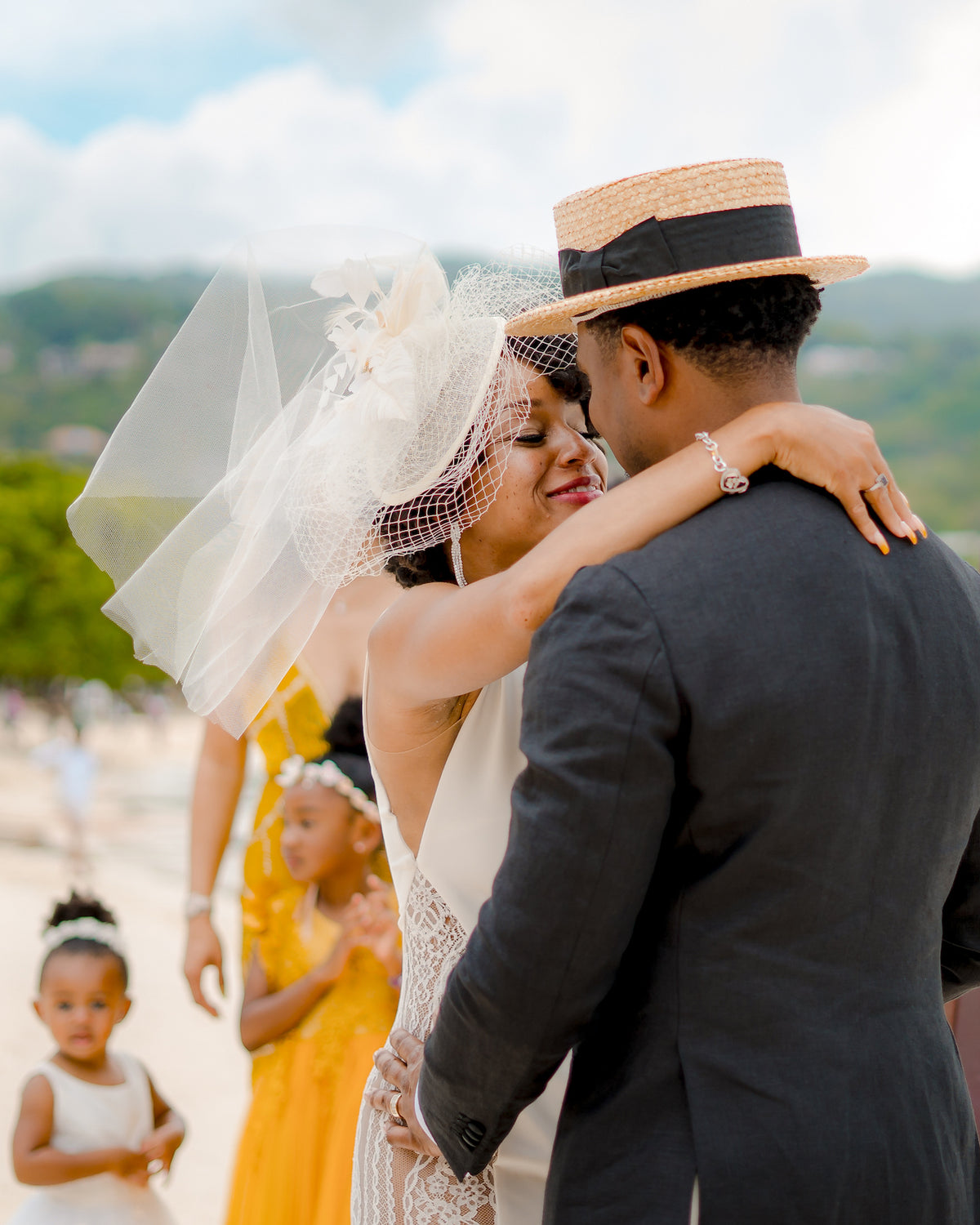 Bride in a birdcage veil embraces her groom in a straw boater hat at a beach wedding in Jamaica