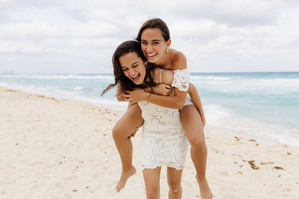 Sisters in white lace dresses laugh during a piggyback on the shoreline at Kempinski Hotel Cancún