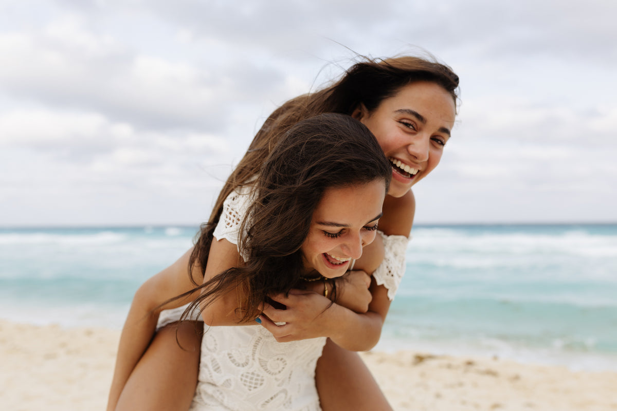 Teen girl gives her laughing sister a piggyback ride on the beach during a lifestyle photoshoot at Kempinski Hotel Cancún