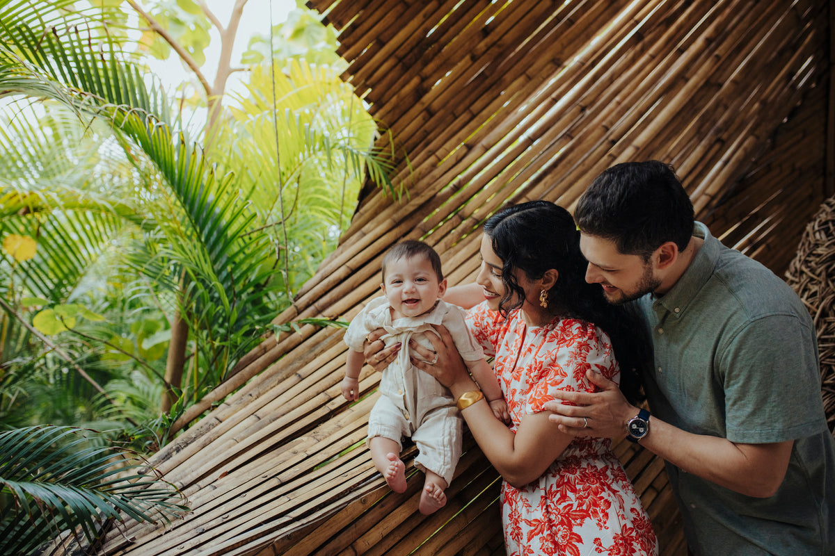 A woman and man smile at a laughing baby inside a bamboo structure surrounded by tropical palms