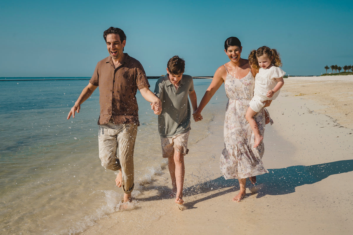 Family of four walking barefoot through shallow water on a sandy beach, hands joined, on vacation