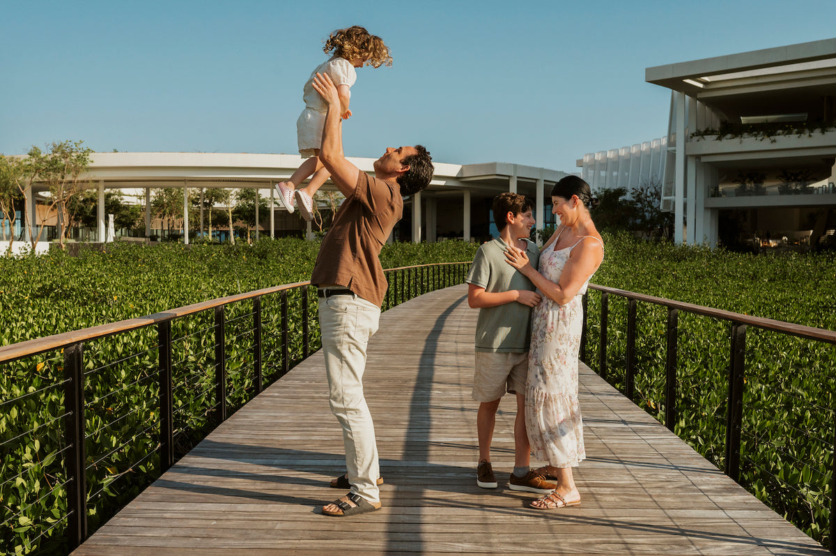 A man lifts a young child overhead on a wooden boardwalk while a woman and boy embrace nearby
