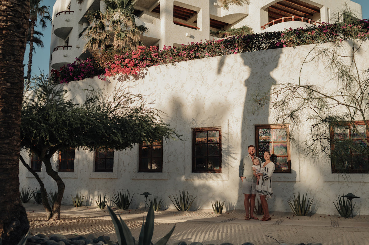A couple holding a baby stands before a white resort building with bougainvillea and agave plants