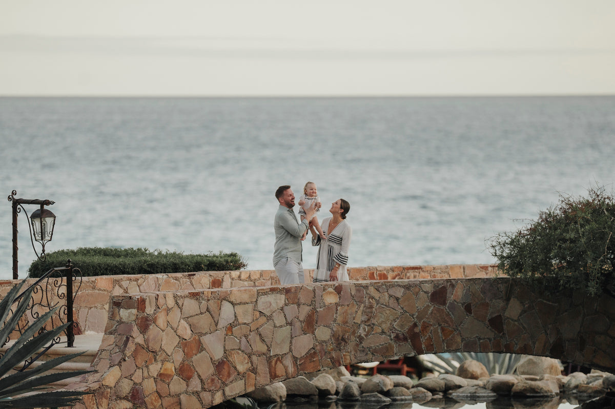 A man holds a toddler up while a woman smiles beside them on a stone bridge overlooking the ocean