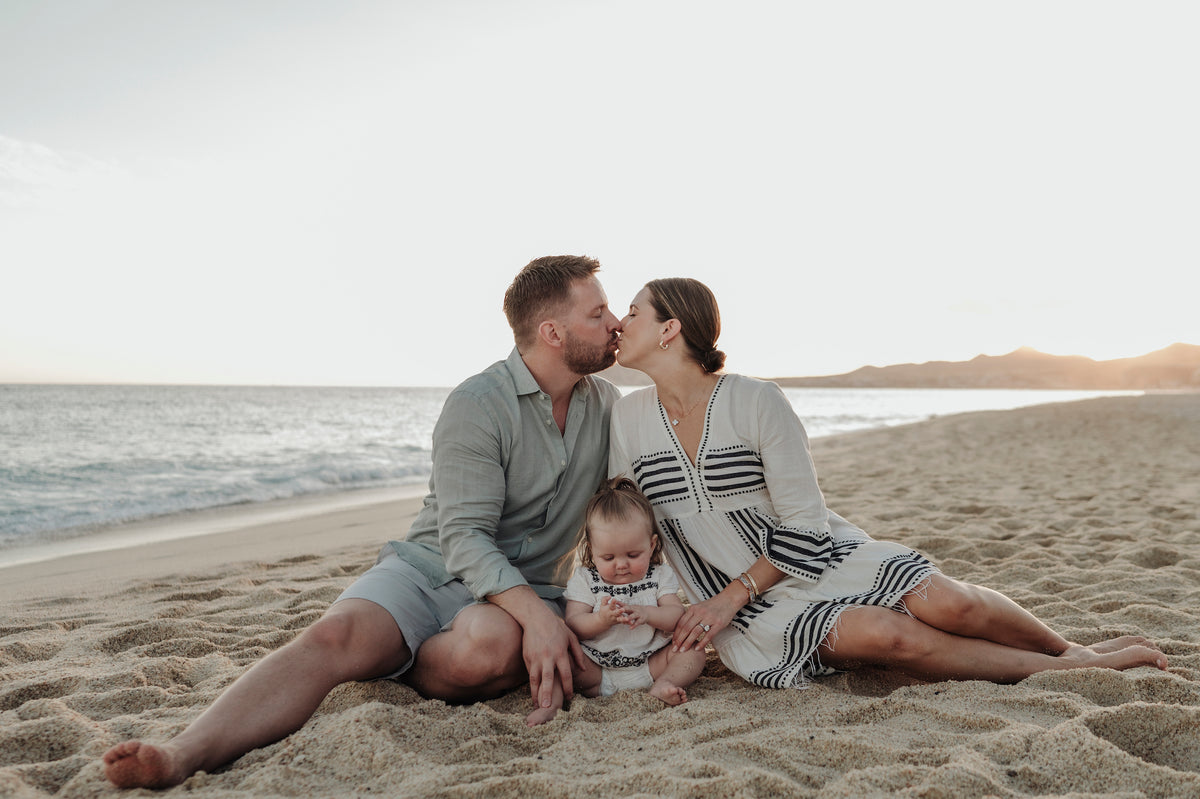A man and woman kiss while sitting on the sand with a toddler between them, ocean behind them