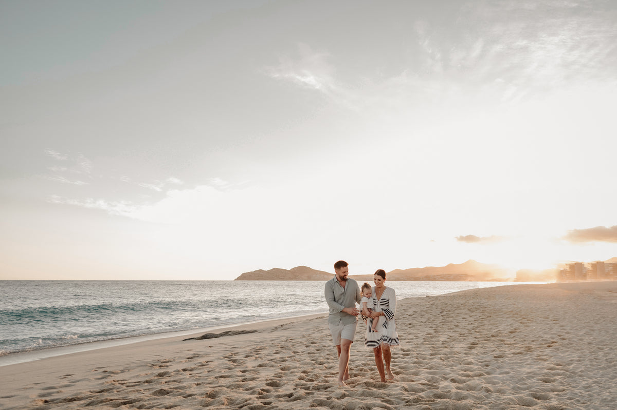 A man, woman, and toddler walk along a sandy beach at sunset with hills in the background