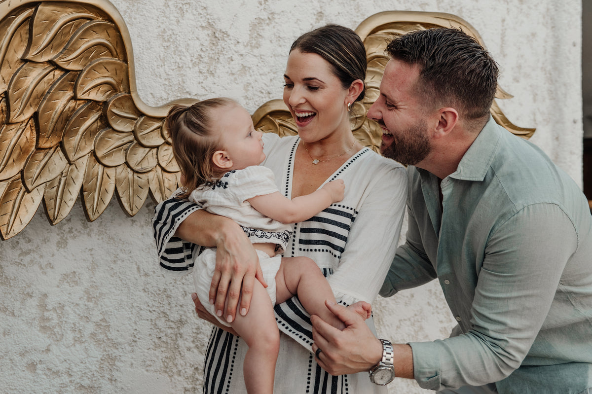 A woman holds a toddler while a man leans in, all laughing beside a gold wing sculpture on a stucco wall
