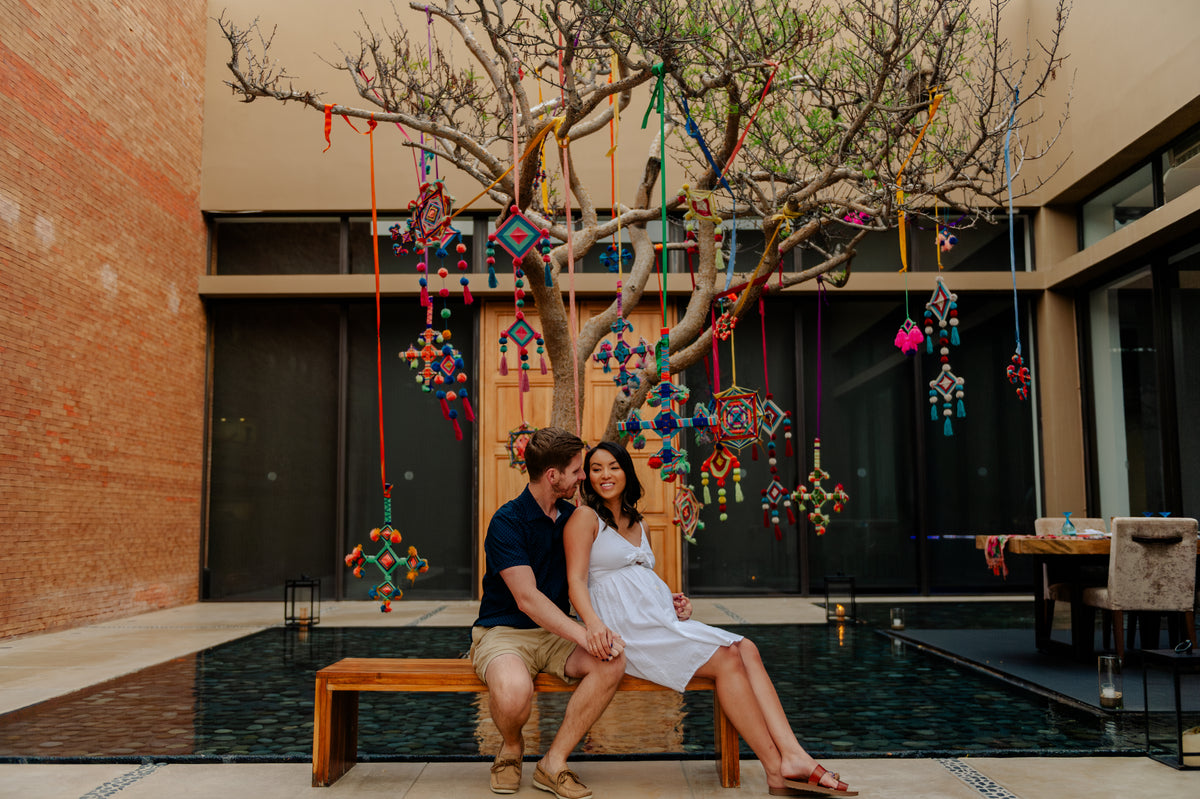 Couple sitting on a bench beneath a tree hung with colorful Mexican pom-pom ornaments in a resort courtyard