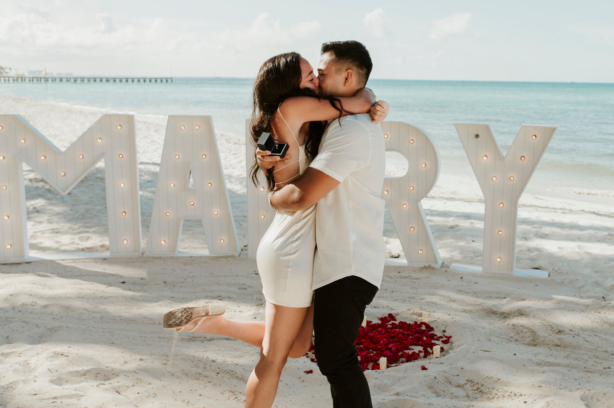 A couple embraces and kisses on a Cancún beach beside large "MARRY" letters at Grand Fiesta Americana Coral Beach