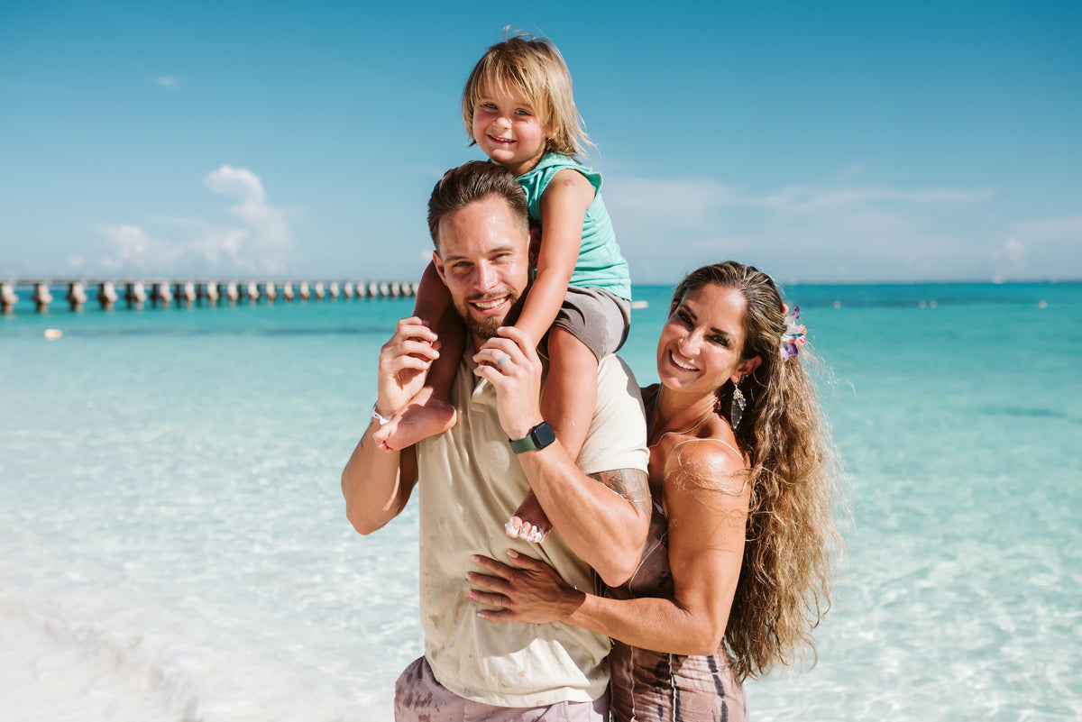 A man carries a child on his shoulders beside a woman on the beach at Grand Fiesta Americana Coral Beach Cancún