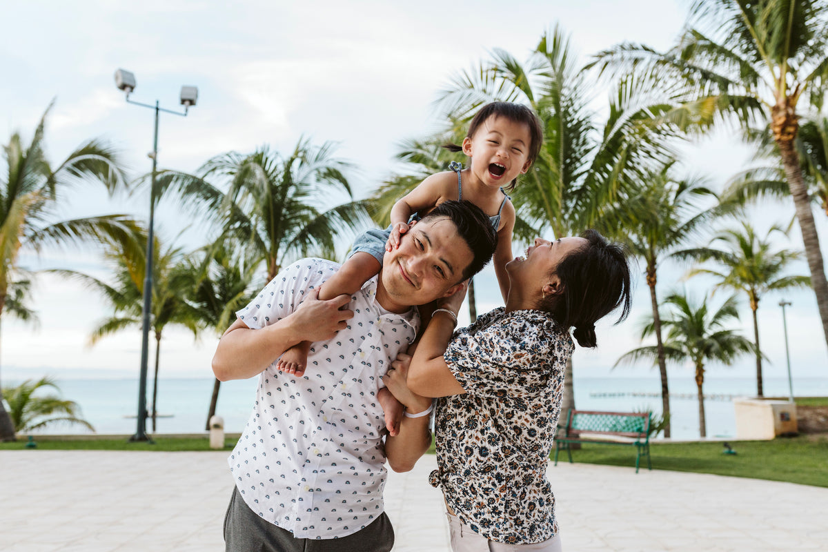 A man carries a laughing child on his shoulders beside a woman, palm trees and ocean behind them at Grand Fiesta Americana Coral Beach Cancún
